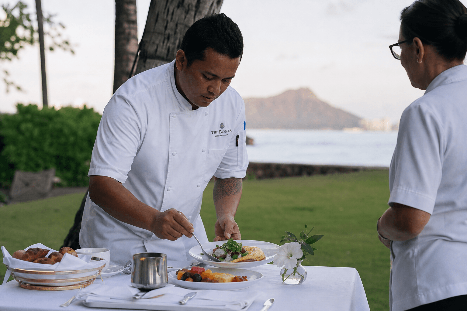 A chef plating recovery brunch fruit and eggs on a Kahala lawn table in morning light, Diamond Head faint.